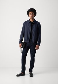 Veste de bombardier marine avec fermeture éclair à l'avant, associée à une chemise noire, un pantalon sombre et des chaussures noires à enfiler, tenant un petit sac noir.