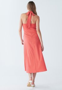 Naine kannab varrukateta korallivärvi keskmise pikkusega kleiti, millel on suur lips kaelal ja valged kontsaga sandaalid, vaatega selja poole.