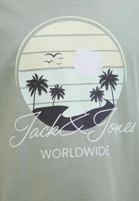 Camiseta verde claro con un gráfico circular que presenta un atardecer, palmeras y el texto "Jack & Jones WORLDWIDE" en blanco.