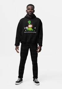 Czarna bluza z kapturem z kolorową grafiką leprechauna i napisem "Szczęśliwego Dnia Picia!", w połączeniu z czarnymi spodniami w szachownicę i sneakersami.