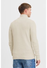 Pulover tricotat de culoare crem, cu guler înalt, tiv cu caneluri și mâneci lungi. Material texturat și o croială lejeră.