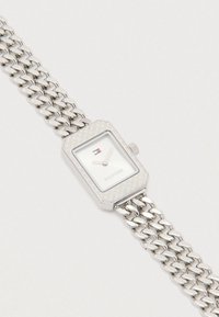 Srebrny prostokątny zegarek z teksturowaną obręczą, białą tarczą i minimalistycznymi wskazówkami. Posiada masywny srebrny bransoletę w formie łańcuszka.