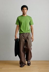 Πράσινο κοντομάνικο μπλουζάκι, καφέ παντελόνι φαρδιάς γραμμής και καφέ παπούτσια. Κρατάει μαύρο σακάκι. Ομαλή υφή υφάσματος, casual εφαρμογή.