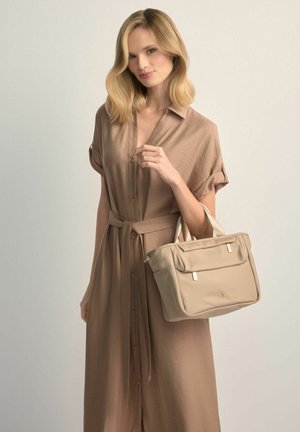 Kobieta o blond włosach do ramion, ubrana w beżową sukienkę z guzikiem, trzymająca dużą beżową torebkę z dwoma uchwytami.