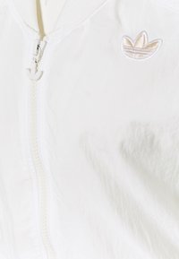 Biała kurtka zapinana na zamek z logo trefoil Adidas na piersi oraz uchwytem do zamka w kształcie trefoila.