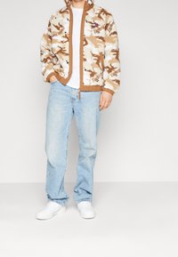 Giacca in pile con zip in camouflage crema e marrone, con polsini e orlo a coste; indossata sopra una maglietta bianca e abbinata a jeans blu chiaro e scarpe bianche.