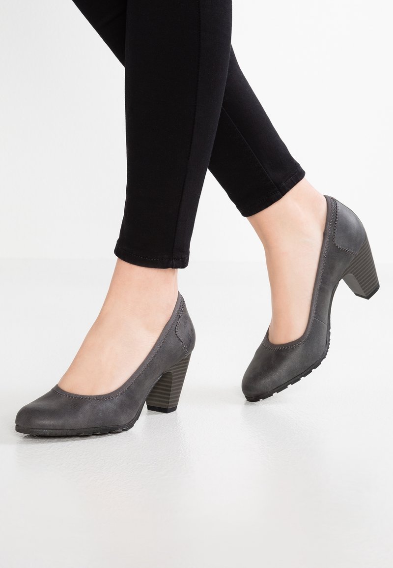 Personne portant un pantalon noir et des chaussures à talons blocs fermées gris foncé, debout sur un sol blanc avec un talon levé.