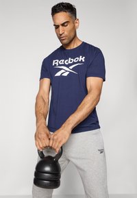 Navy Reebok t-shirt with large white logo, paired with light gray sweatpants, holding a black kettlebell, focused expression.