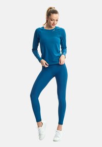 Kobieta w niebieskiej bluzce z długim rękawem i legginsach oraz białych trampkach stoi na jednolitym białym tle, poprawiając swoją koszulę.