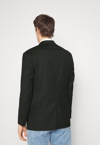 Schwarze Hautton Blazer mit glatter Textur, mit einer Schlitze und drei Knopfmanschetten, getragen über hellblauen Jeans.