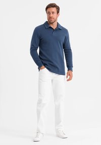 Camisa de polo de manga larga en azul marino con cuello, con puños acanalados y un dobladillo recto, acompañada de pantalones blancos y zapatillas blancas.