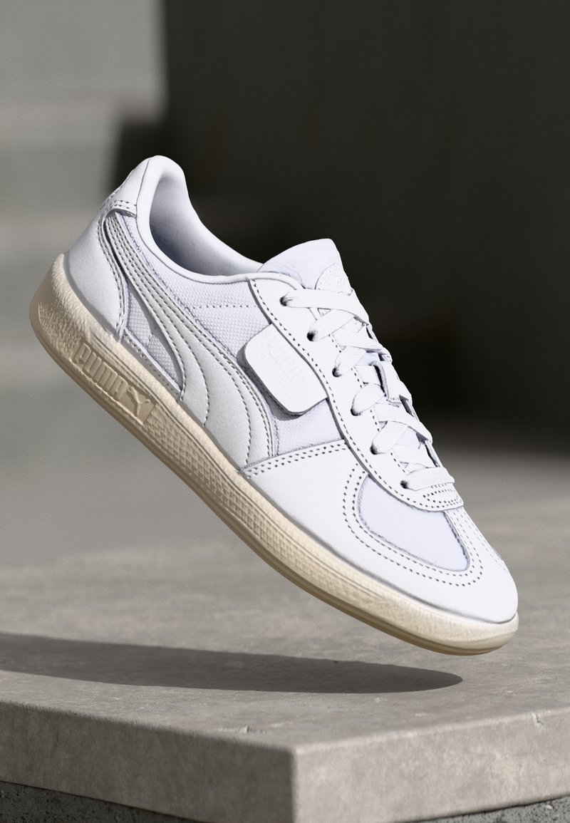 Chaussure de sport blanche en cuir texturé et panneaux en mesh, avec un patch logo en relief, des lacets ronds et une semelle en caoutchouc beige.