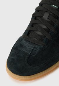 Dark green suede sneaker with a rounded toe, black laces, and a contrasting gum rubber sole. Smooth texture with stitched accents.