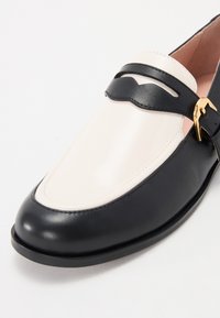 Black and white leather loafer with a shiny finish, featuring a black strap and gold buckle accent, rounded toe, and smooth texture.