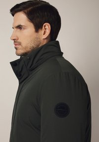 Mężczyzna z krótkimi ciemnymi włosami, ubrany w ciemnozieloną kurtkę z wysokim kołnierzem i okrągłą łatą na rękawie, zwrócony w lewo na jednolitym tle.