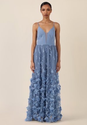 Frau in einem bodenlangen, staubblauen Kleid mit Spaghettiträgern, tiefem V-Ausschnitt und 3D-Blütenapplikationen auf dem transparenten Rock.