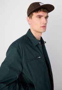 Donkergroene zip-up jas met een kraag en een ritszak op de borst. Gecombineerd met een bruine pet. De stof lijkt glad en lichtgewicht.