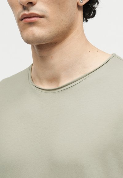 Κοντινό πλάνο στο κάτω μέρος του προσώπου και τον λαιμό ενός ατόμου που φοράει ανοιχτό πράσινο μπλουζάκι με στρογγυλή λαιμόκοψη και ένα μικρό στρογγυλό ασημένιο σκουλαρίκι.