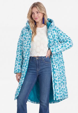 Junge Frau, die einen langen Steppmantel mit blauem Blumenmuster, ein weißes gehäkeltes Oberteil, blaue Jeans und blaue Quastenohrringe trägt, steht mit einer Hand in der Hüfte.
