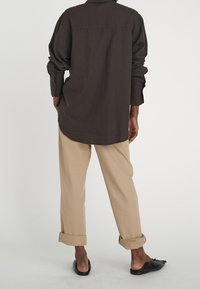 Brown long-sleeve shirt with relaxed fit and button details, paired with rolled-up beige trousers and black slip-on shoes.