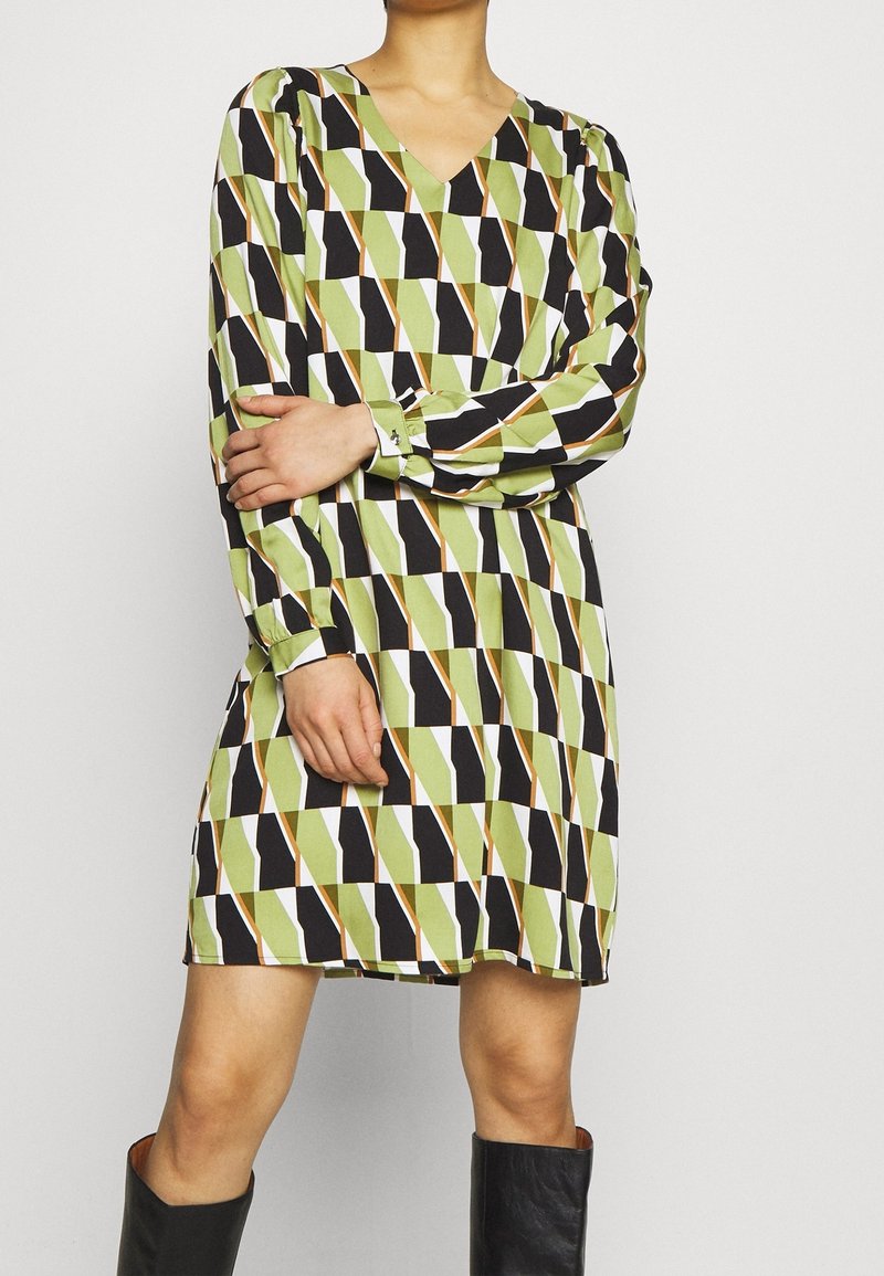 Personne portant une robe courte à manches longues avec un motif géométrique vert, noir et blanc, associée à des bottes noires mi-mollet.