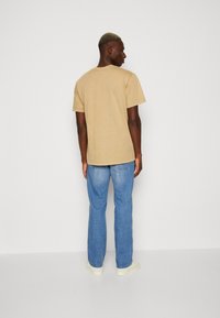 Tan cotton t-shirt with a relaxed fit, round neckline, and short sleeves, paired with blue straight-leg jeans and white sneakers.