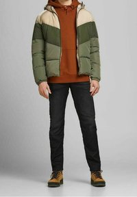 Padded jacket w oliwkowym i beżowym kolorze z kapturem, noszony na pomarańczowej bluzie z kapturem. Zestawiony z czarnymi dżinsami i brązowymi butami trekkingowymi.