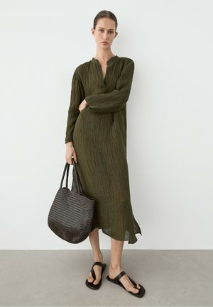 Donna che indossa un lungo vestito verde oliva in tessuto strutturato con maniche lunghe, tiene una grande borsa intrecciata scura e indossa sandali neri.