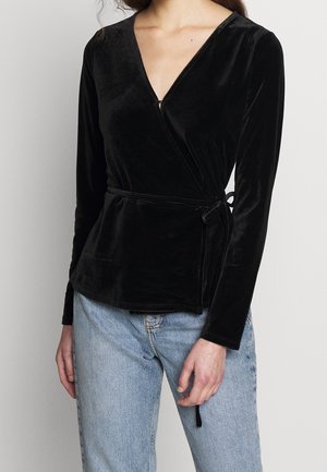 Kvinde iført en sort velour omslagstop med lange ærmer og lyseblå jeans, stående mod en ensfarvet lys baggrund.