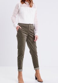 Biała fakturowana bluzka z falbankami, zestawiona z oliwkowozielonymi spodniami ze sztruksu oraz beżowymi botkami za kostkę na słupku.