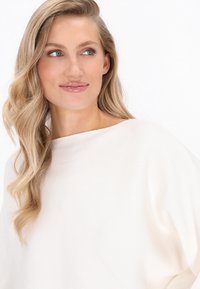 Haut couleur crème avec un large col bateau et une texture légèrement côtelée, présentant un drapé doux et une coupe décontractée. Cheveux longs et ondulés visibles.
