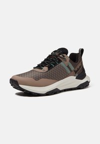 Sneaker Timberland marrone e nero con tessuto strutturato, strisce verde chiaro, suola bianca spessa e suola esterna nera robusta.