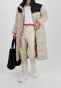 Abrigo largo acolchado con un diseño en bloques de color beige y negro, que presenta un cierre de cremallera, cuello alto y bolsillos laterales. Lleva puesto sobre pantalones crema.