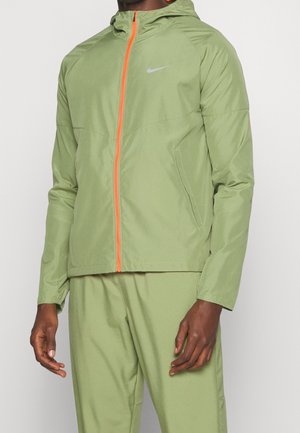 Person trägt einen hellgrünen Nike-Trainingsanzug mit Kapuzenjacke, die einen orangefarbenen Reißverschluss und Seitentaschen aufweist.