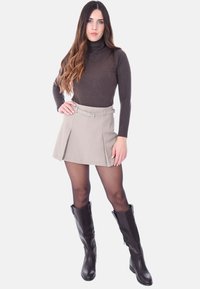 Maglione dolcevita marrone, gonna a trapezio beige con cintura, stivali neri al ginocchio, tessuto liscio e design aderente, posa in piedi con le mani sui fianchi.