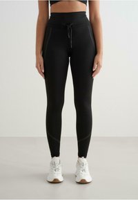 Leggings negre cu talie înaltă și șnur, cu cusături laterale și un material neted și elastic. Asortate cu adidași albi.