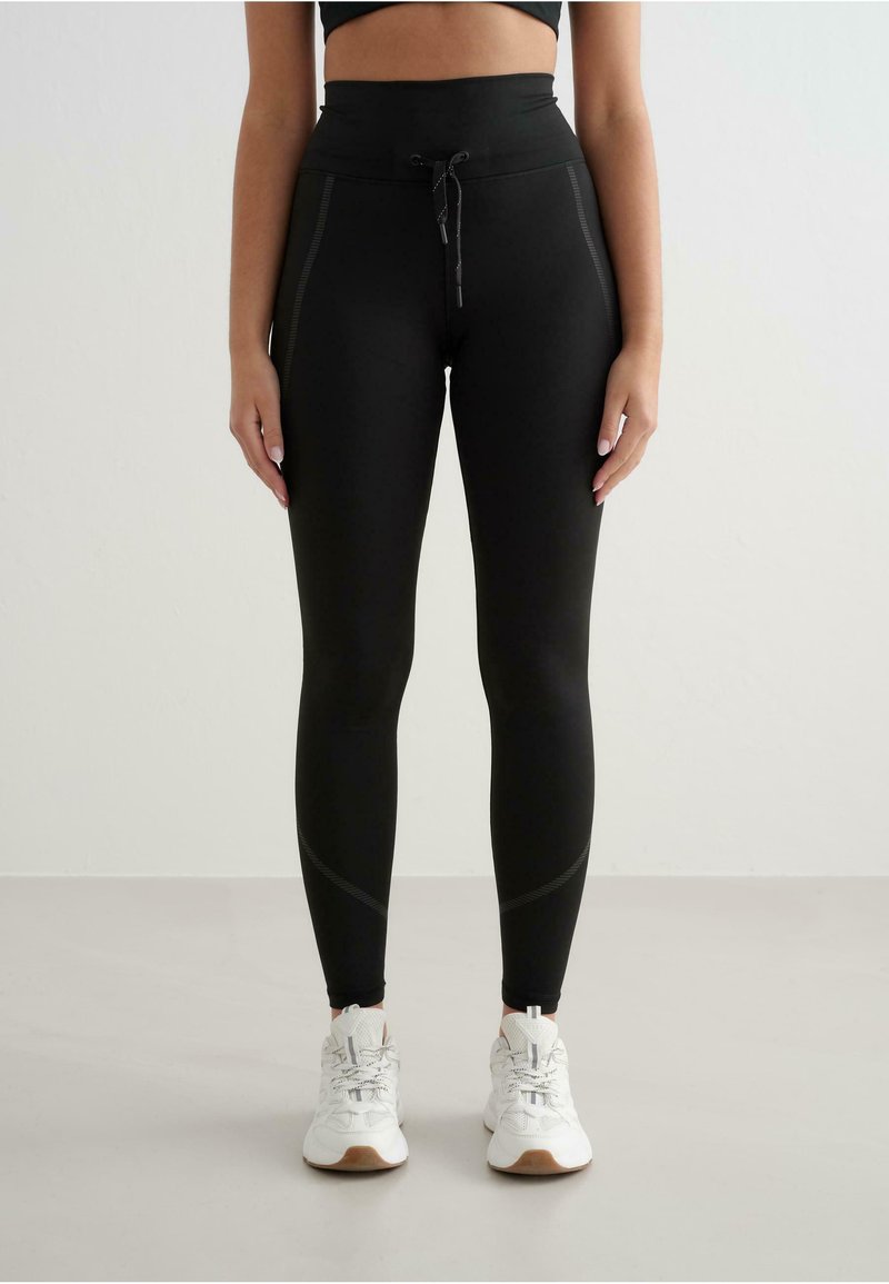 Leggings negre cu talie înaltă și șnur, cu cusături laterale și un material neted și elastic. Asortate cu adidași albi.