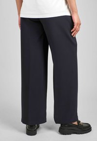 Hosen mit weitem Bein aus dunkelblauem Stoff, die über eine glatte Textur und einen entspannten Schnitt verfügen. Kombiniert mit schwarzen Slippern für einen vollständigen Look.