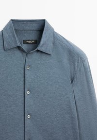 Camicetta button-up blu scuro con colletto, tessuto morbido e strutturato, cinque bottoni sul davanti e un'etichetta del marchio all'interno del colletto.