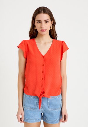 Giovane donna con lunghi capelli castani che indossa una camicetta arancione vivace con bottoni, annodata in vita, e pantaloncini di jeans azzurri.