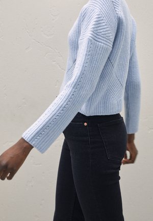 Lyseblå ribbestrikket sweater med diagonale strikkemønstre og lange ærmer, parret med sorte jeans med en lille kobberknapdetalje.