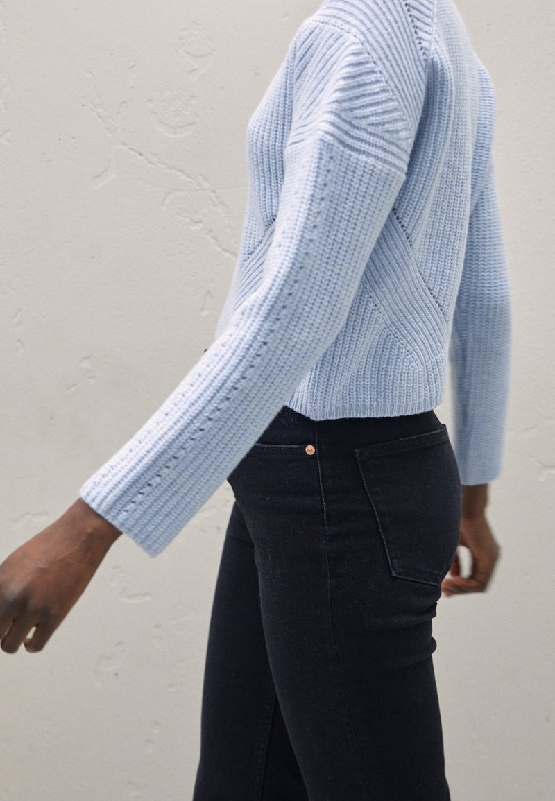Maglione azzurro chiaro a coste con motivi a maglia diagonale e maniche lunghe, abbinato a jeans neri con un piccolo dettaglio in rame sui bottoni.