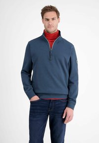 Niebieski sweter z zamkiem błyskawicznym do połowy, z prążkowanymi mankietami i dołem, noszony na czerwonym golfie. Zawiera logo na lewym rękawie. Dżinsy.