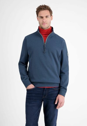 Blaues Viertelzip-Sweatshirt mit gerippten Bündchen und Saum, über einem roten Rollkragenpullover getragen. Mit einem Logo auf dem linken Ärmel. Jeans aus Denim.