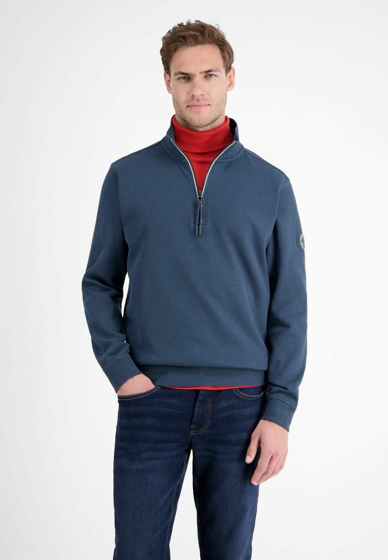 Niebieski sweter z zamkiem błyskawicznym do połowy, z prążkowanymi mankietami i dołem, noszony na czerwonym golfie. Zawiera logo na lewym rękawie. Dżinsy.