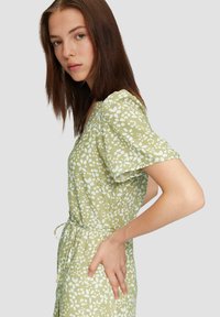 Zielona sukienka w kwiaty z białymi wzorami liści. Posiada dekolt w kształcie litery V, krótkie bufiaste rękawy oraz wiązany pasek w talii. Materiał wydaje się lekki i miękki.