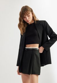 Blazer nero sopra un top corto nero aderente e una gonna nera plissettata a vita alta. Presenta una fibbia argentata e una superficie in tessuto liscia.