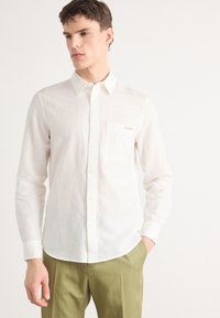Camicia bianca button-up in tessuto leggero con finitura testurizzata, dotata di taschino sul petto e dettaglio logo discreto. Abbinata a pantaloni verdi.