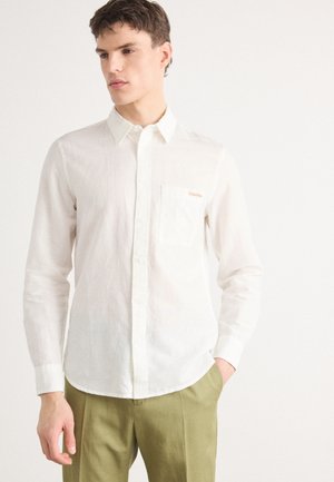 Chemise blanche à manches longues en tissu léger avec une finition texturée, dotée d'une poche poitrine et d'un logo discret. Associée à un pantalon vert.