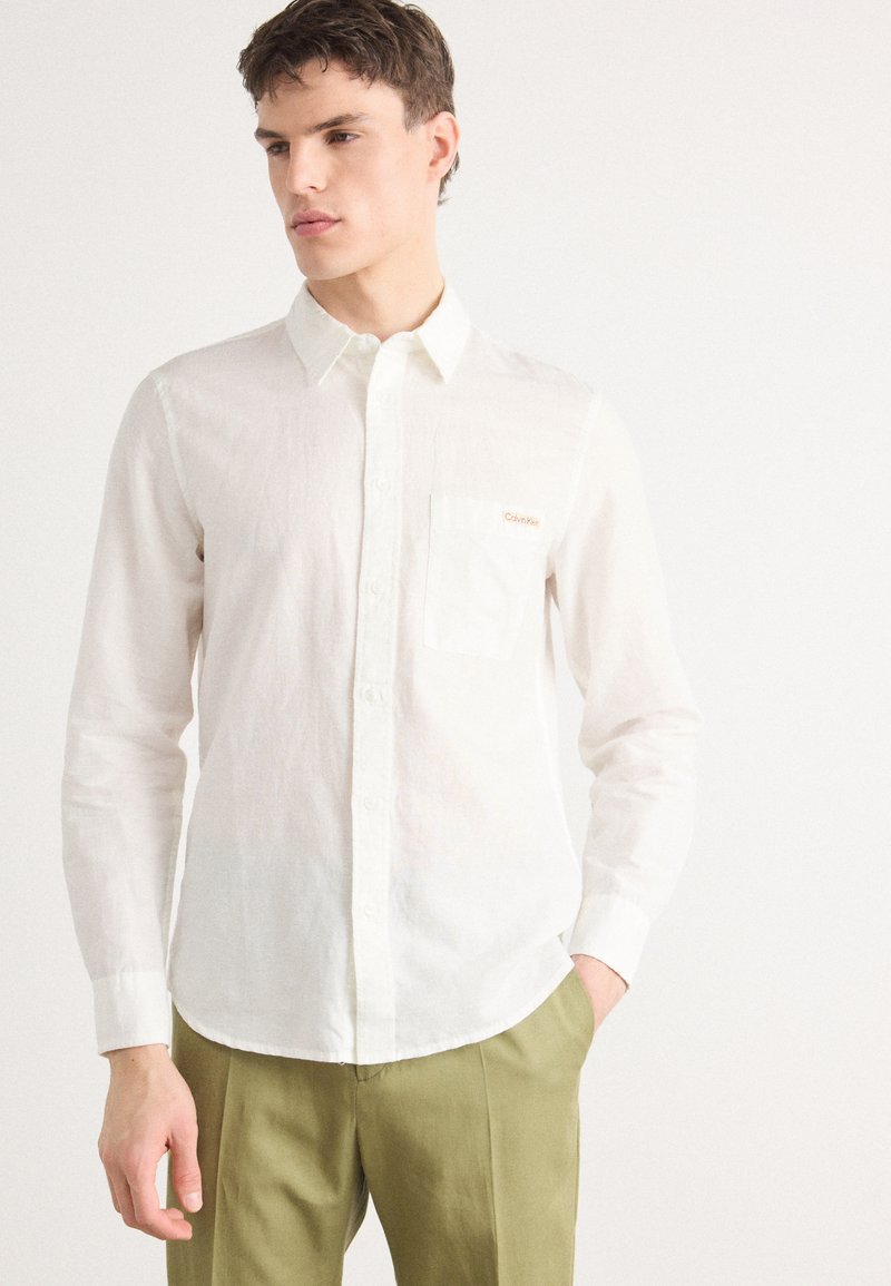 Camicia bianca button-up in tessuto leggero con finitura testurizzata, dotata di taschino sul petto e dettaglio logo discreto. Abbinata a pantaloni verdi.
